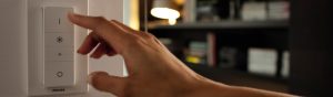 ”Hand justerar Philips smarta LED-dimmerströmbrytare för att styra den omgivande belysningen i ett mysigt vardagsrum med bokhyllor.”